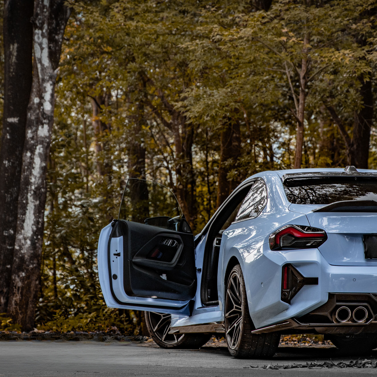 A light blue car with its driver's door open is parked on a forest road, surrounded by tall trees and dense foliage.