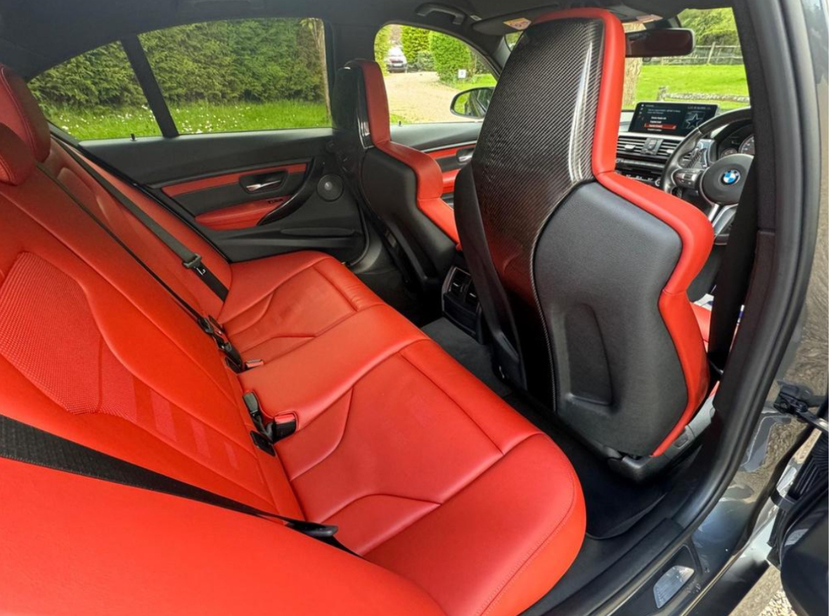Car interior featuring vibrant red leather seats, providing a striking contrast against a sleek black dashboard. The setting is a parked car on a driveway with greenery outside.