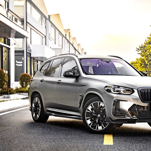 A silver SUV is parked on a city street, surrounded by modern buildings and greenery in an urban setting, with sunlight illuminating the scene.