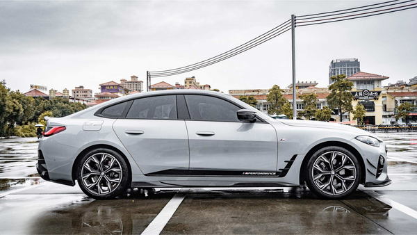 A sleek white car with tinted windows is parked on wet pavement, displaying the text "M PERFORMANCE" on its side. Buildings and trees form the urban background.