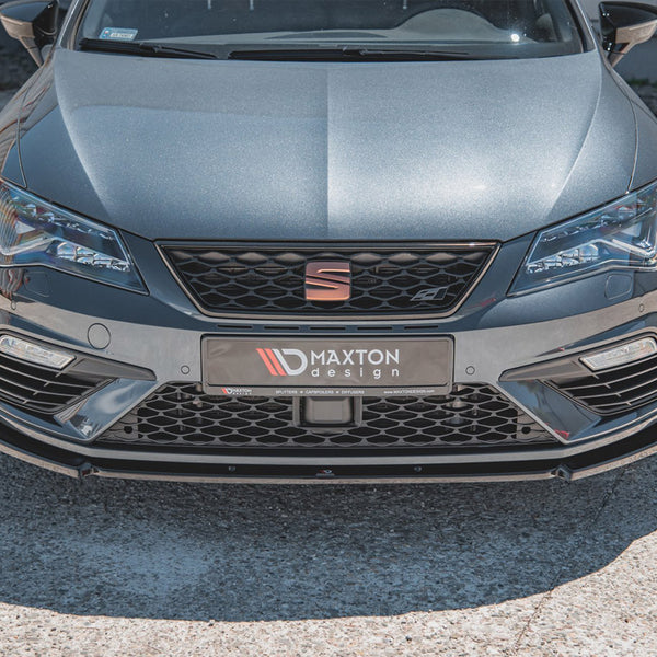 A sleek grey car displays a Maxton Design front bumper with sharp headlights in a sunlit parking area. The front plate reads, “MAXTON design” and includes small text: “SPLITTERS COMPOSITES DIFFUSERS www.MAXTONDESIGN.com.”