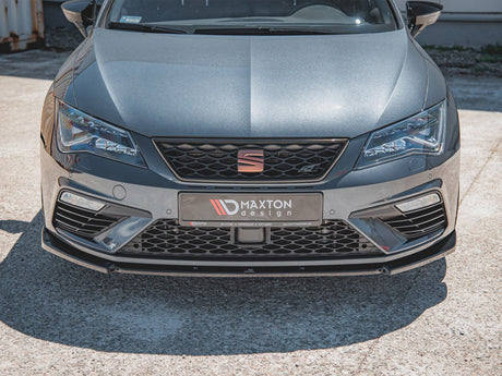 A sleek grey car displays a Maxton Design front bumper with sharp headlights in a sunlit parking area. The front plate reads, “MAXTON design” and includes small text: “SPLITTERS COMPOSITES DIFFUSERS www.MAXTONDESIGN.com.”