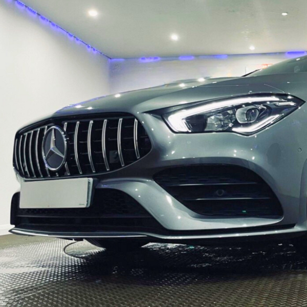A silver car with LED headlights and a distinctive front grille displays a logo, parked indoors on a textured metal floor under bright ceiling lights. No visible text.