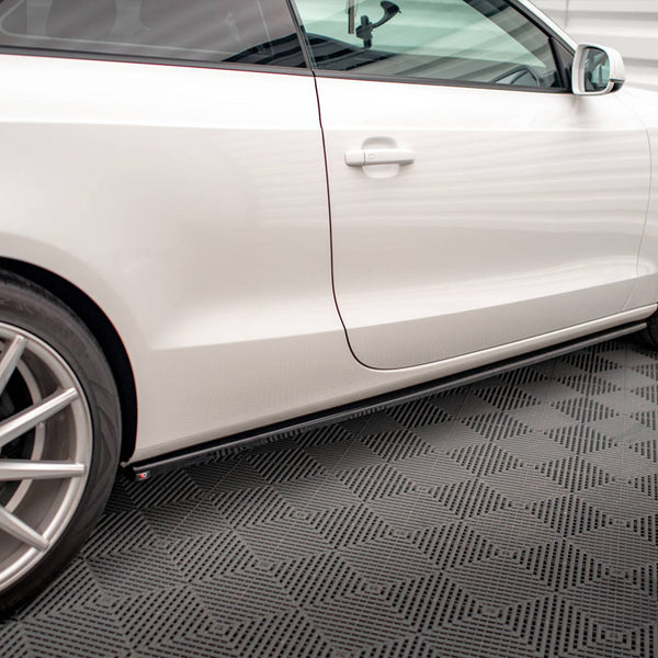 A white car parked on a patterned garage floor, viewed from the side, revealing its sleek design, shiny wheels, and closed door. The setting has a modern and clean appearance.