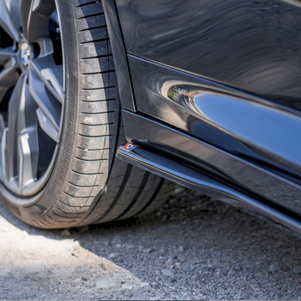 A close-up of a black car's rear wheel on a gravel surface, with a shiny side skirt reflecting nearby surroundings. The wheel features a modern, multi-spoke design.