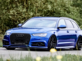 A blue Audi station wagon with black wheels is parked on a road, surrounded by greenery and blurred yellow flowers. The grille features an Audi logo and 