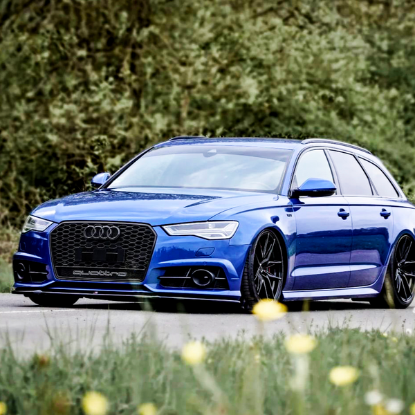 A blue Audi station wagon with black wheels is parked on a road, surrounded by greenery and blurred yellow flowers. The grille features an Audi logo and "quattro" text.