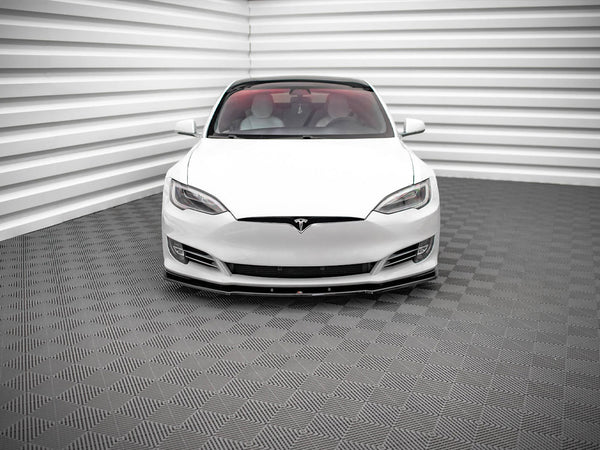 A white car, viewed from the front, is parked in a room with textured, gray-patterned flooring and walls featuring horizontal white panels.