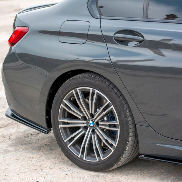 A dark-colored car is parked, showcasing a close-up of its rear wheel and tire on a concrete surface. The car's sleek and polished design is visible in bright daylight.