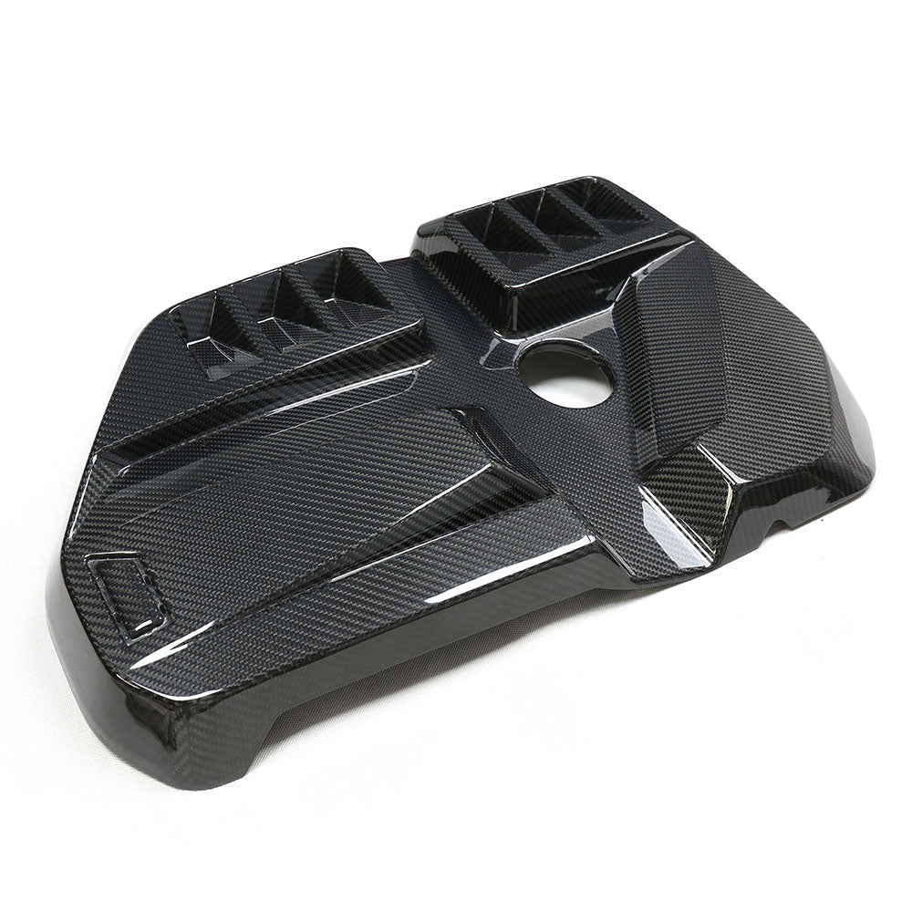 Carbon fiber engine cover with a glossy finish, featuring geometric ridges and a central circular hole, positioned on a white background.