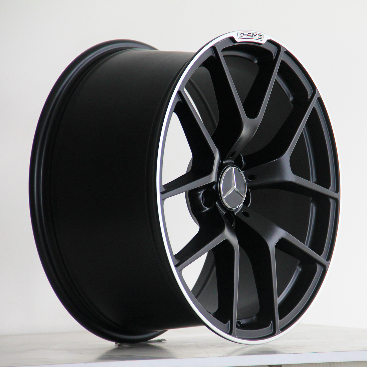 A black alloy wheel with multiple spokes features a Mercedes emblem in the center. It rests on a plain surface. The text "AMG" is visible on the wheel's rim.