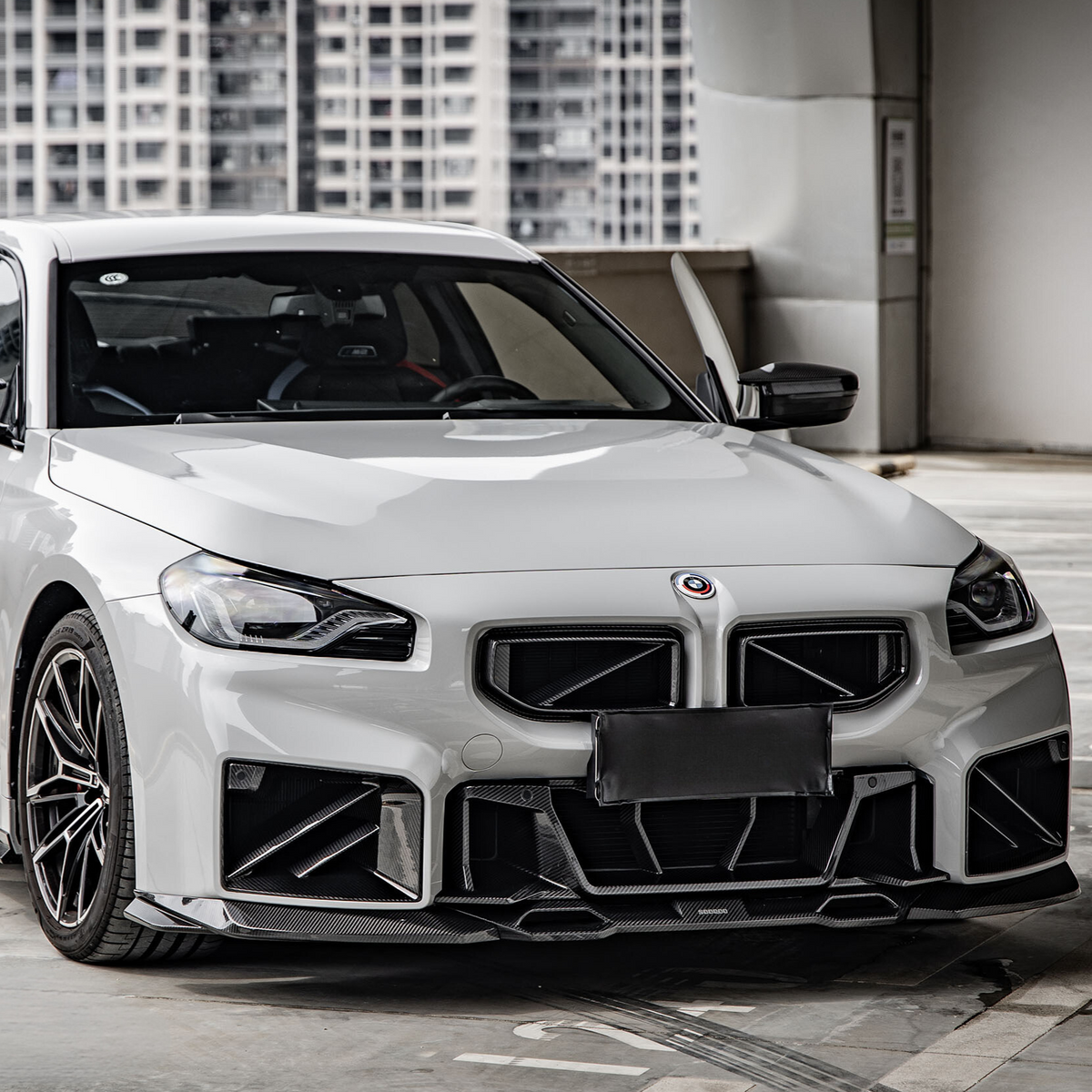 A white, sporty car is parked in a modern urban setting, featuring a bold front grille and sleek design. High-rise buildings are visible in the background.