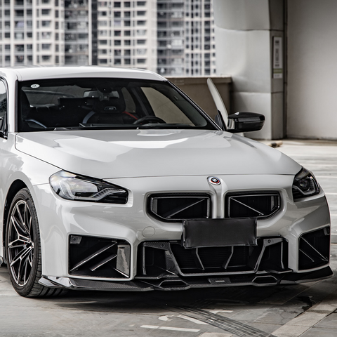 A white, sporty car is parked in a modern urban setting, featuring a bold front grille and sleek design. High-rise buildings are visible in the background.
