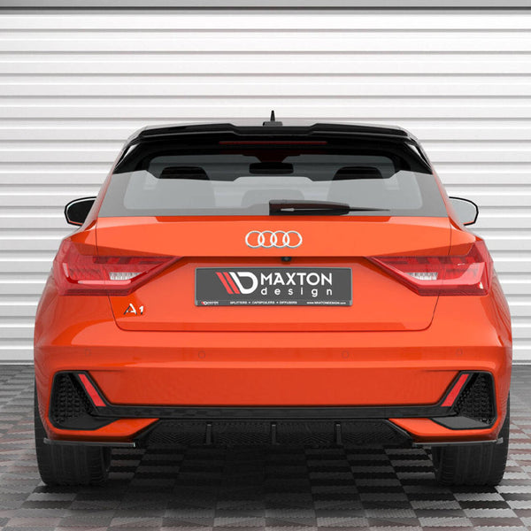 A bright orange car is parked on a checkered floor facing a white garage door. Text on the license plate reads "MAXTON design." The car is labeled "A1" and has Audi's logo.
