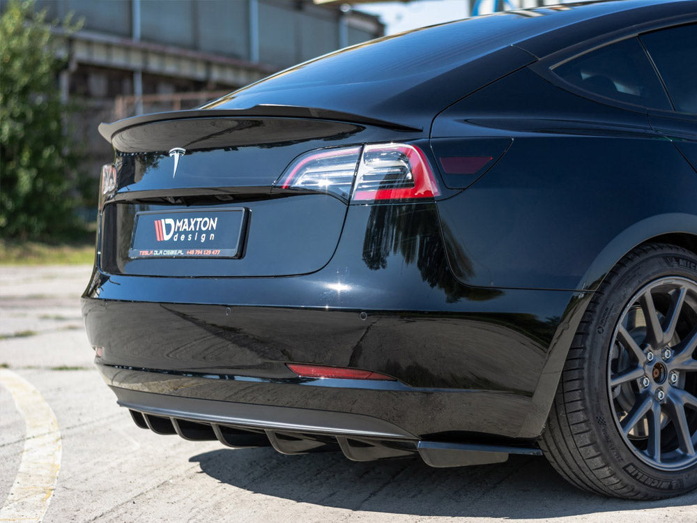 Black car rear with a Maxton Design license plate frame. It features a sleek spoiler and diffuser. The car is parked outdoors on a sunny day beside a building.