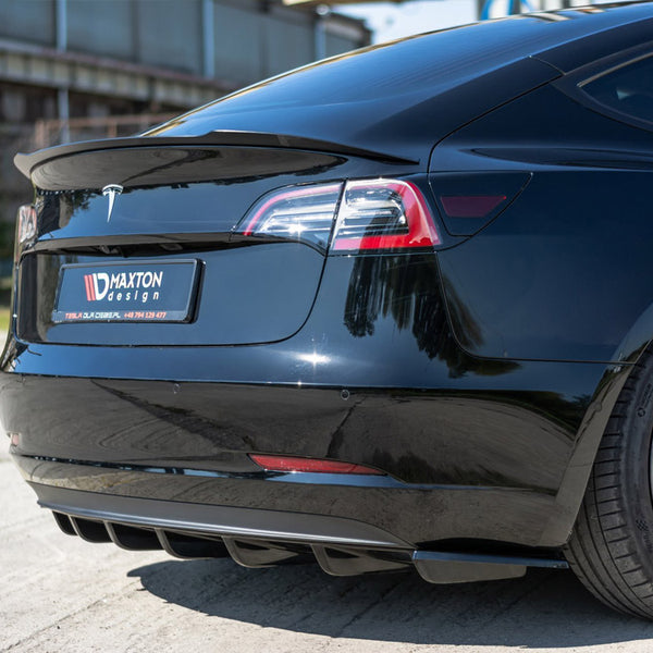 Black car rear with a Maxton Design license plate frame. It features a sleek spoiler and diffuser. The car is parked outdoors on a sunny day beside a building.