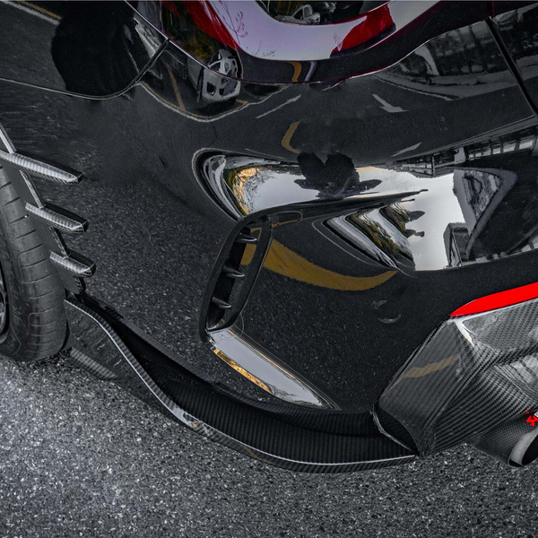 A sleek black sports car's rear is shown with dual exhaust pipes, carbon fiber detailing, and a red accent. It's parked on a textured, grey asphalt surface.