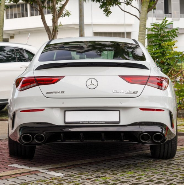 A white Mercedes-Benz CLA 45 S, shown from the rear, displays AMG and model badges. It's parked on a brick surface with trees and a building in the background.