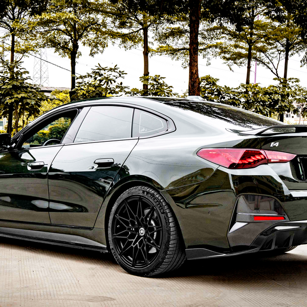 A sleek, dark-colored car is parked on a concrete surface. It features prominent, stylized tail lights and black rims. Trees and shrubs provide a backdrop, suggesting an outdoor setting.