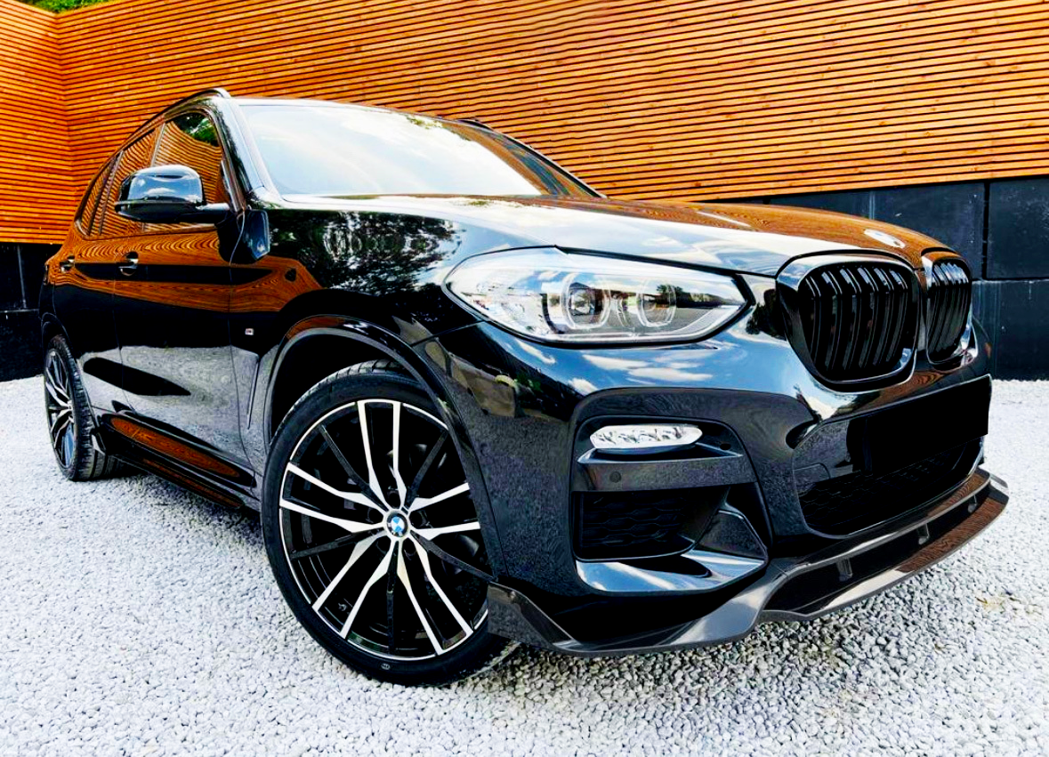 A sleek black car with stylish alloy wheels parked on gravel, positioned in front of a modern wooden slat wall.