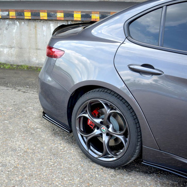 A gray car is parked, showing its rear wheel with red brake calipers. The setting is an outdoor paved area adjacent to a concrete barrier with black and yellow markings.