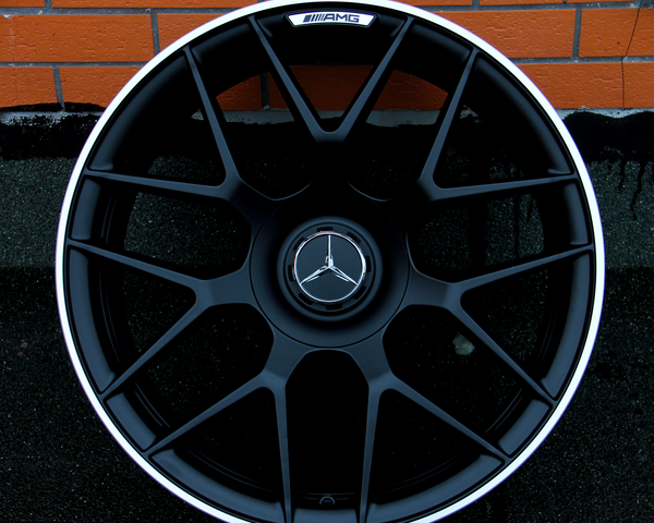 A black alloy wheel with a Mercedes-Benz logo at the center rests against a textured orange brick wall. The top of the wheel features the text "AMG."