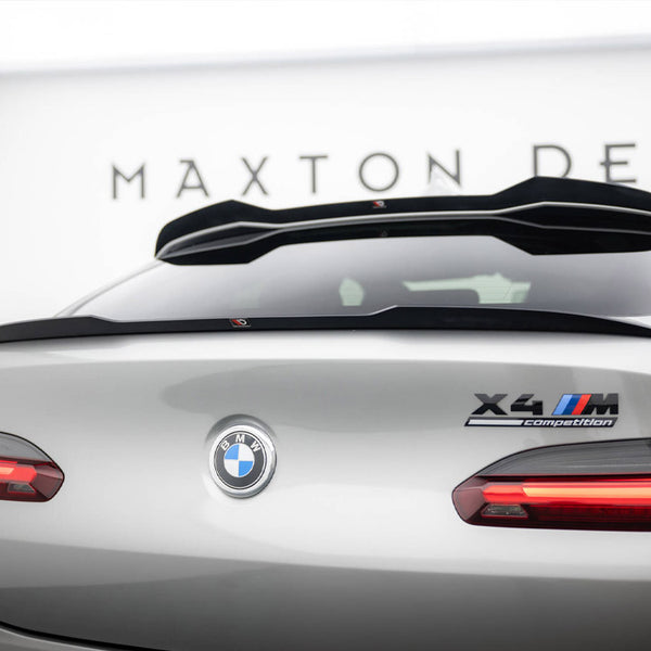 Silver BMW car viewed from the rear, featuring "X4 M Competition" badge. In a showroom setting with "MAXTON DESIGN" on the wall, taillights are illuminated.