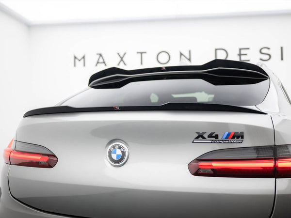 Silver BMW car viewed from the rear, featuring "X4 M Competition" badge. In a showroom setting with "MAXTON DESIGN" on the wall, taillights are illuminated.