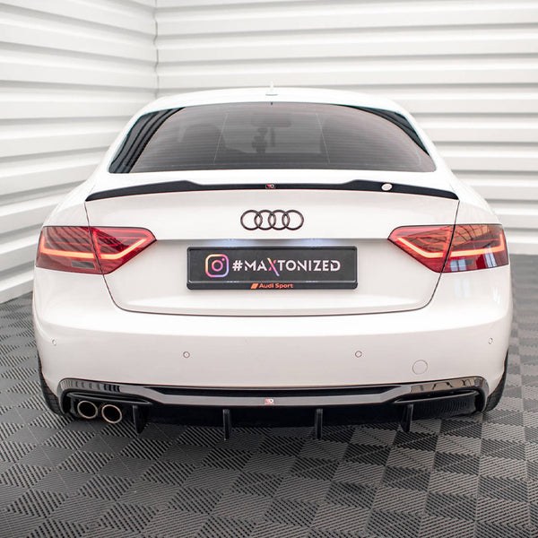 A white Audi car is stationary in a room with white, ribbed walls and checkered flooring. The rear license plate displays "#MAXTONIZED" with Instagram and Audi Sport logos.