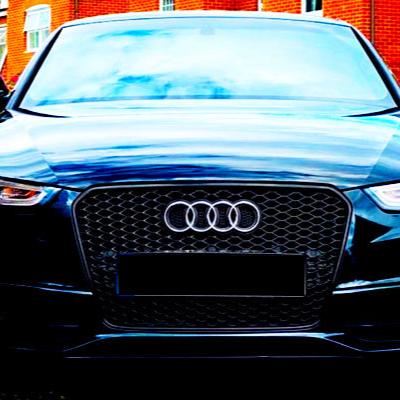A black Audi car is stationary, facing forward, with its grille and logo prominently visible. It's parked in front of a red brick building with reflective glass windows.