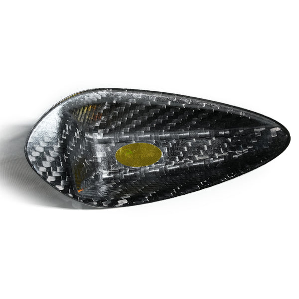 Triangular carbon fiber bowl with a glossy weave texture rests on a white surface. A small, gold oval sticker with text is inside, adding contrast to the dark material.