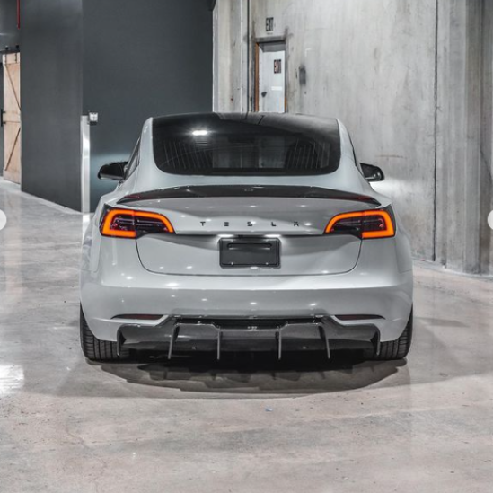 A silver Tesla car is parked in a concrete garage. Its taillights are illuminated, and "TESLA" is visible on the back. An "EXIT" sign appears on a door in the background.