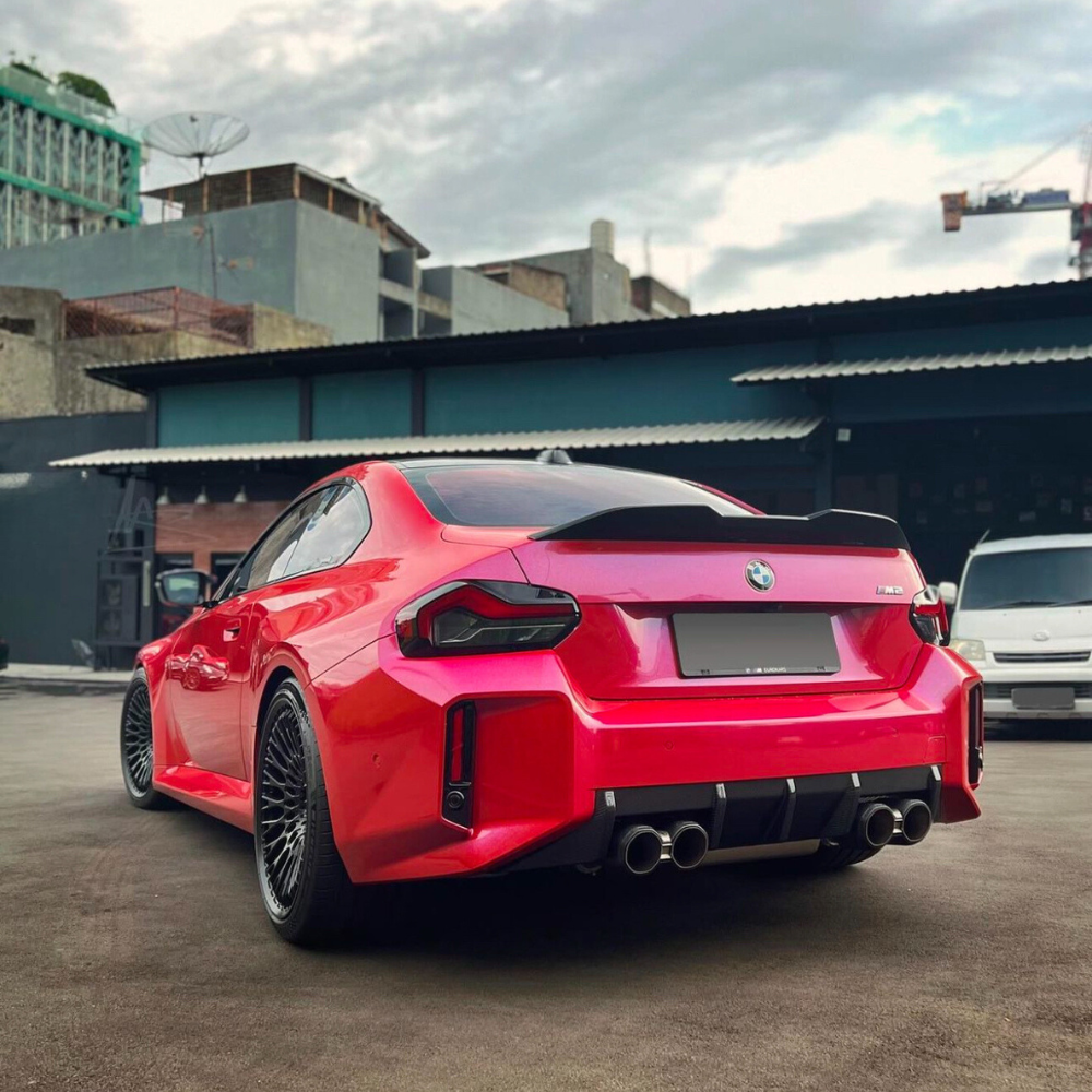 A bright red car is parked, showcasing its sleek design with a sporty spoiler and dual exhausts. It sits in an urban setting with industrial buildings and a cloudy sky.