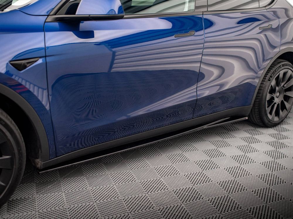 A blue car parked indoors with wavy patterns reflecting on its glossy surface, sitting on a textured, checkered floor. Black wheels and sleek design enhance its modern appearance.
