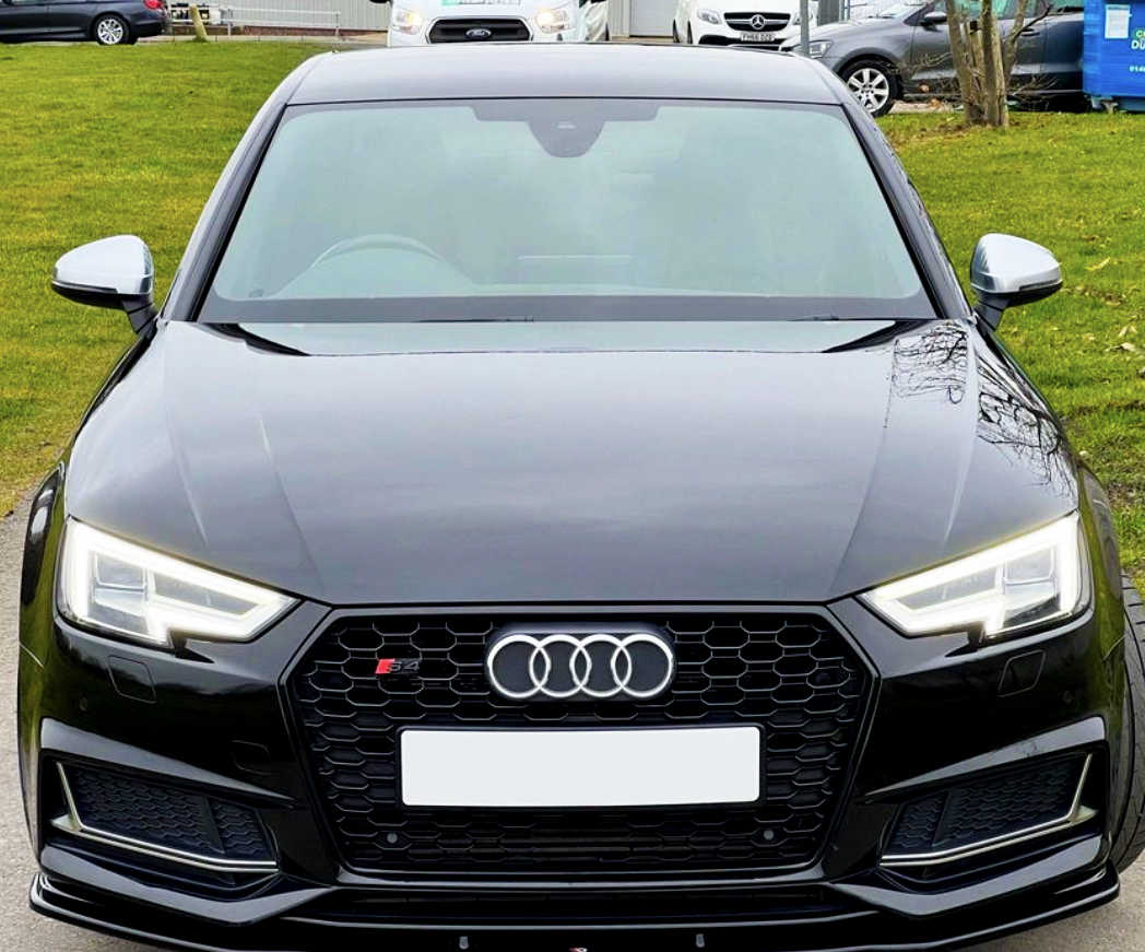 A black Audi S4 is parked on a road, viewed from the front with its headlights on. In the background, other cars and greenery are visible.