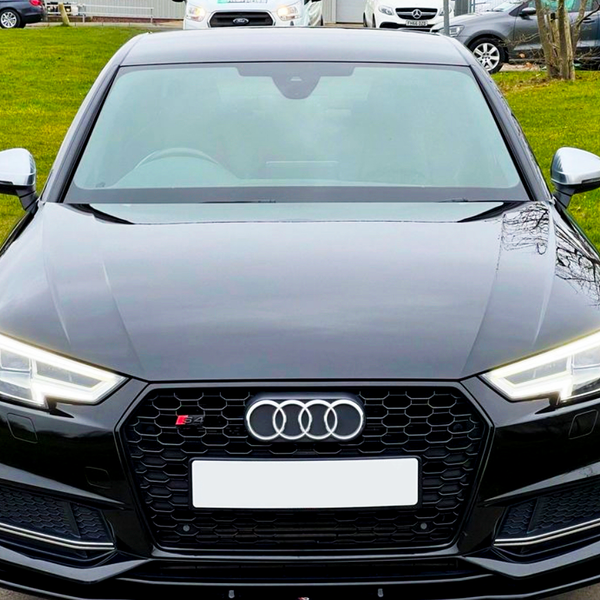A black Audi S4 is parked on a road, viewed from the front with its headlights on. In the background, other cars and greenery are visible.