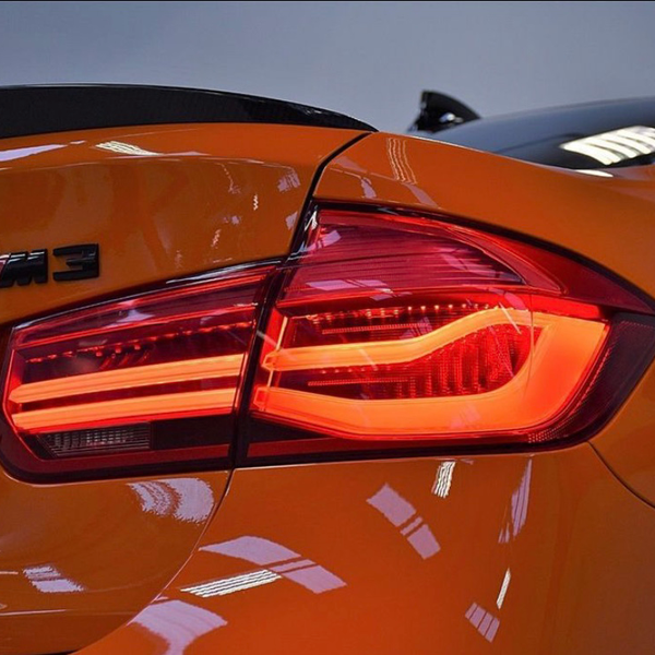A brightly lit rear taillight on an orange sports car, with the model badge "M3" visible, set against a neutral, indoor background.