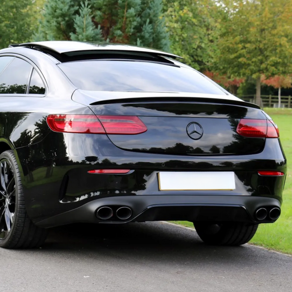 A black car with a sunroof open is parked on a road, surrounded by grass and trees. The rear features dual exhaust pipes and a Mercedes-Benz logo. License plate is blank.