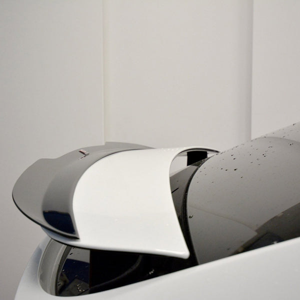 A white car's retractable spoiler is partially deployed, with water droplets on its surface, set against a plain indoor background.