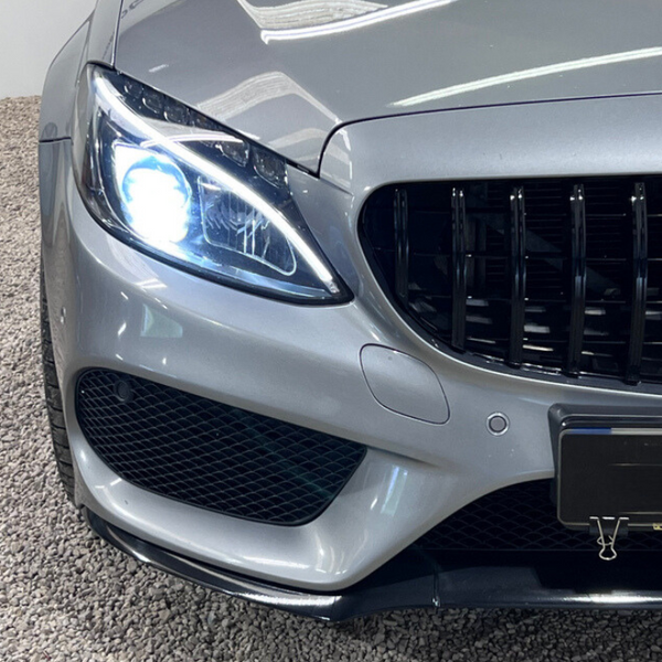 A silver car with illuminated left headlight and black grille is parked on gravel. The grille features vertical bars and a circular emblem, while a binder clip secures the front license plate.