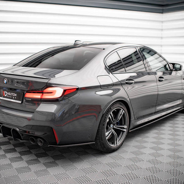 A sleek, gray sedan is parked in a modern, checkered-floor showroom. Its rear features distinctive taillights and dual exhaust pipes. The license plate reads "MAXTON design."