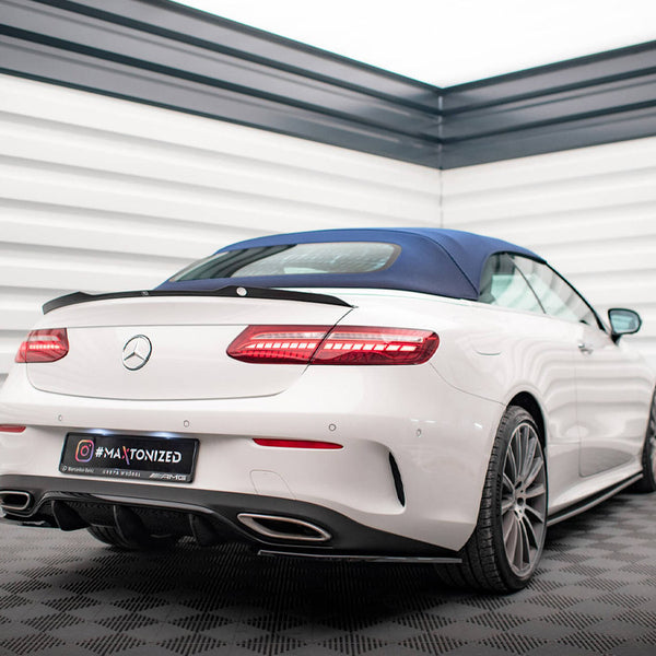 A white convertible car is parked in a modern showroom with white paneled walls. It features a blue roof and visible AMG branding. Text on license plate reads: "#MAXTONIZED."