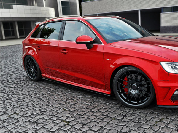 A red car with black wheels is parked on cobbled pavement, surrounded by modern, gray buildings in an urban setting.