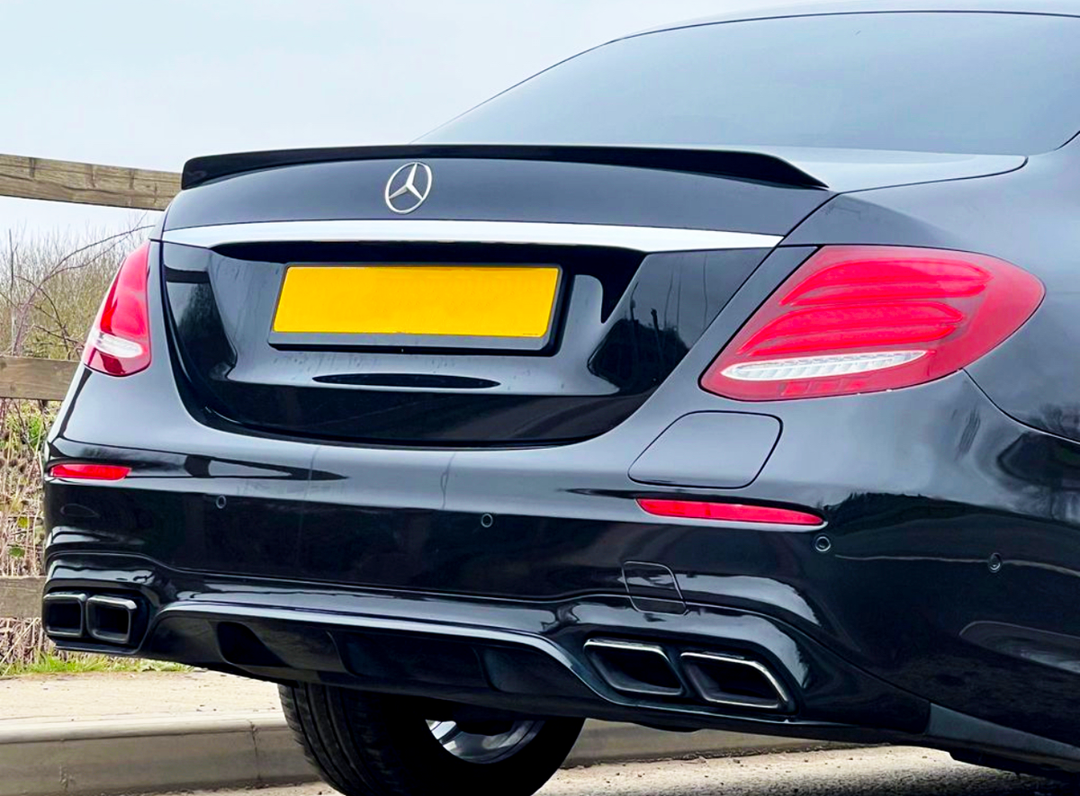 A black car with a visible rear, featuring Mercedes emblem, tinted windows, and dual exhausts, is parked on a road near wooden fencing and greenery. The license plate is yellow.