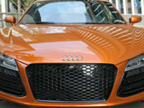 An orange Audi car, parked on a city street, displays a honeycomb grille and Audi emblem, with modern buildings reflecting on its shiny surface.