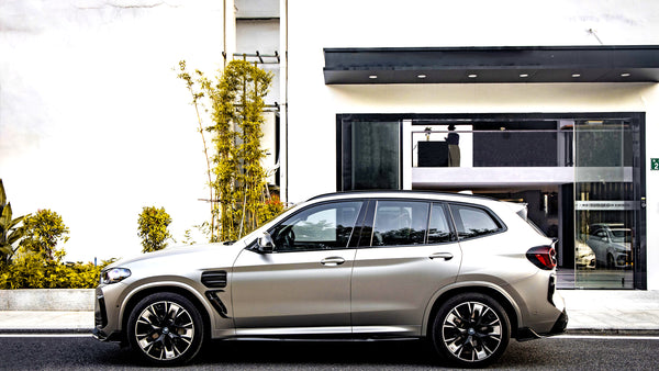 A silver SUV is parked beside a modern building with a black entrance. Yellow-green plants line the sidewalk. Reflections show other vehicles and signage: "INSTITUTO DE DRY CARBON".