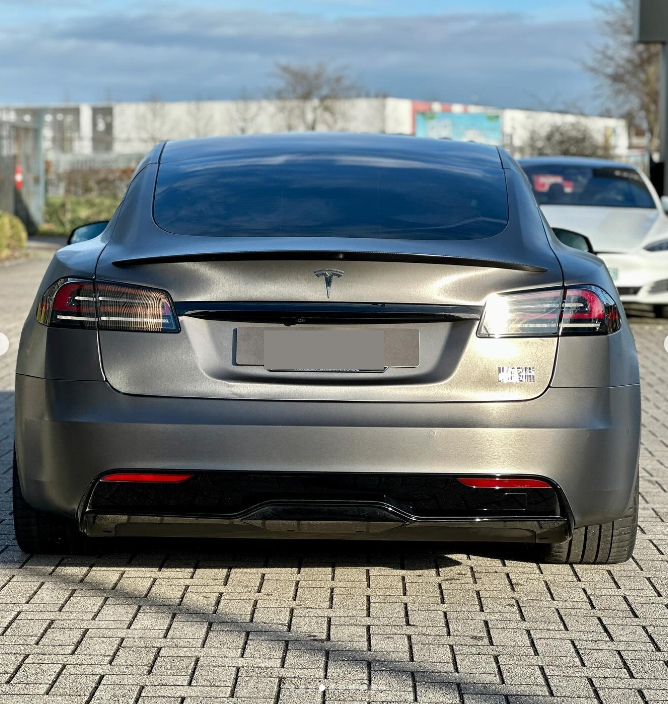 A gray Tesla Model S is parked on a brick driveway, surrounded by other vehicles. The license plate is blurred, and there are trees and buildings in the background.