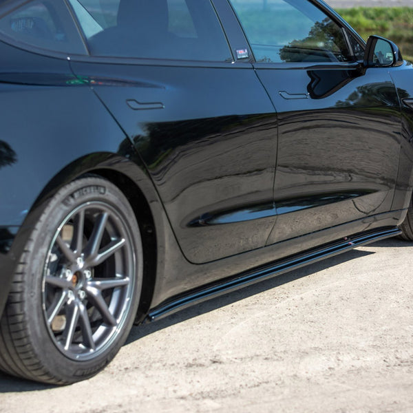 A sleek black car is parked on a sandy path beside a green area. The car's reflective surface mirrors the surrounding trees and sky.