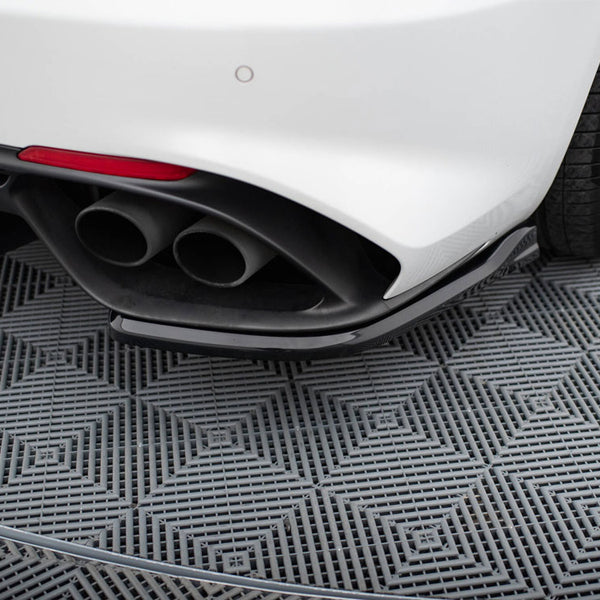 Car exhaust pipes protrude from the rear of a white vehicle, parked on a gray, textured, diamond-patterned surface, with a visible rear tire and partial red reflector.