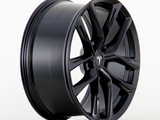 A black, multi-spoke alloy wheel displaying a Tesla logo, stands upright on a plain white background, showcasing a sleek design with a polished finish.